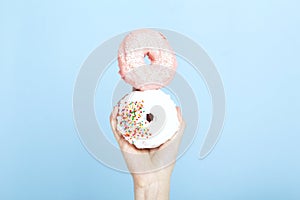 Woman hand holds two donuts with pink and white  icing and multicolored sugar pastry topping, blue background