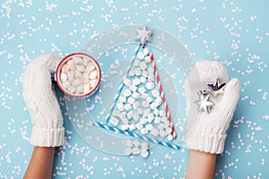 Woman hand in mitten hold cup of hot cocoa or chocolate and christmas fir tree made of marshmallow on turquoise winter background