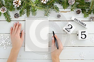 Woman hand with pen and blank notebook. Christmas decoration Fir tree, Pine cones and perpetual calendar 25 december on