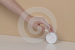 Woman hand is picking up the blank cosmetic packaging mockup: white bottle on beige background.