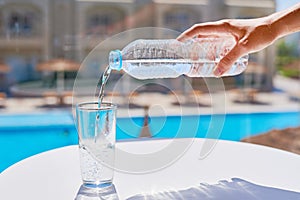 Woman hand pouring water in a glass from plastic bottle agains swimming pool background