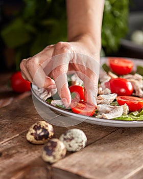 Woman hand put tomato cherry to a ceramic plate of fresh salad with organic ingredients on a wooden table. Healthy diet