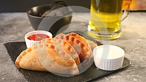 Woman hand puts glass of beer on the concrete table with Mexican fried meat pies empanadas cheburek on the black plate.