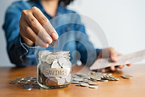 The woman hand is putting a coin in a glass bottle and a pile of coins on a brown wooden table,Investment business, retirement,