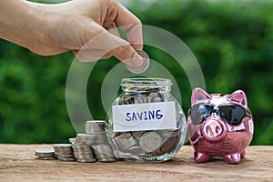 Woman hand putting coin in glass jar bottle with stack of coins