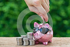 Woman hand putting coin in glossy pink piggy bank with stack of