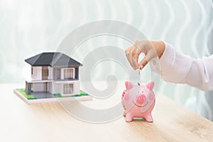 Woman hand putting a coin into a piggy bank on wooden desk - save money to buy new house concept