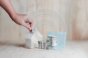 Woman hand putting the coin in to wooden house piggy bank financial metaphor saving money for buy home concept on wooden table