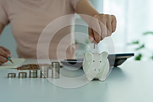 Woman hand is putting coins in a piggy bank with piles of coins placed next to them to save money as expenses and investments. The