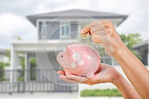 Woman hand putting money coin in to piggy for saving money wealth and financial concept with home in background, Saving for buy