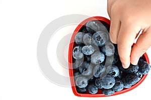 Woman hand with red nails picking fresh ripe blueberry from a red heart bowl, on white table