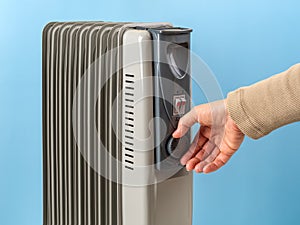 Woman hand rotates the thermostat knob of oil filled electric heater against blue background. Portable household appliance for