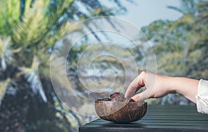 Woman hand with royal dates fruit in a bowl of coconut on a table in a palm tree grove.