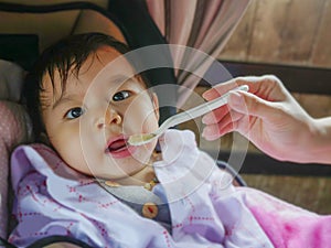 Woman hand with spoon feeding her daughter, a sweet and adorable beautiful Asian Chinese baby girl 7 or 8 months old sitting at