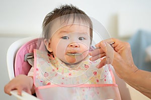 woman hand with spoon feeding puree to happy and adorable baby girl in a bib sitting on eating chair in child healthy nutrition