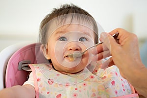 woman hand with spoon feeding puree to happy and adorable baby girl in a bib sitting on eating chair in child healthy nutrition