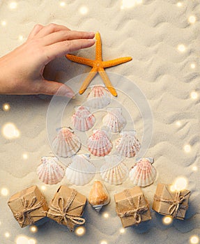 A woman hand straightens a starfish on a Christmas tree made of seashells on the sand. Travel season to sunny countries and the