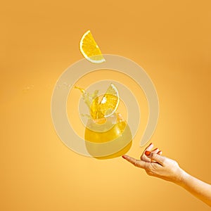 Woman hand support fly glass of orange drink with splash, juice slice orange falling in glass. Summer art food concept