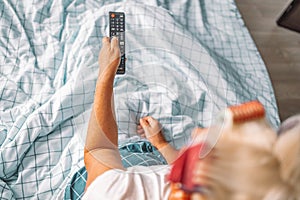 Woman hand switches channels on the TV while sitting in bed under a white blanket. View from above