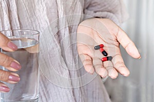 Woman hand taking antibiotic pills red and black capsules with glass of water