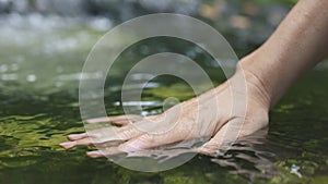 Woman hand touches water in transparent mountain river, waterfall on background. Slow motion 4K shot of hand on surface