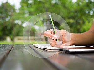 Woman hand use green pencil writing on white notebook on table in green natural park. Think out side the box and generate idea in