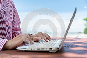 Woman hand using laptop to work study on work desk with clean nature beach outdoor background. Business, financial concept