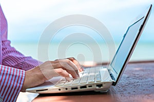 Woman hand using laptop to work study on work desk with clean nature beach outdoor background. Business, financial concept