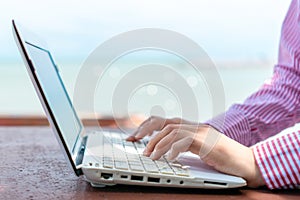 Woman hand using laptop to work study on work desk with clean nature beach outdoor background. Business, financial concept