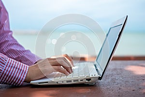 Woman hand using laptop to work study on work desk with clean nature beach outdoor background. Business, financial concept