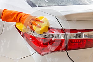 Woman hand wearing orange gloves with yellow sponge washing taillight modern car or cleaning automobile.