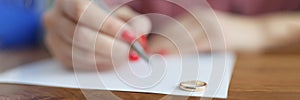 Woman hand writing declaration of divorce on background of wedding rings closeup