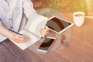 Woman hand writing notebook and phone, tablet on table in garden