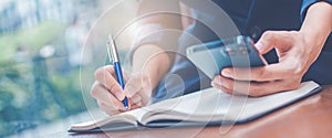 Woman hand writing on a notepad with a pen and she is holding a cell phone in the office