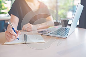 Woman hand is writing on a notepad with a pen.On the table, there is a laptop computer and a cup of coffee