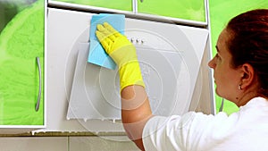 Woman hand in yellow household gloves spray detergent to surface of kitchen hood closeup. Housewife carry out wet