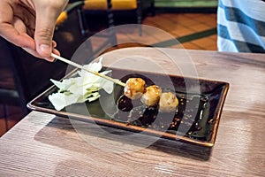 Woman handing holding a stick of grilled meatball in spicy dipping sauce served with fresh cabbage on brown porcelain plate