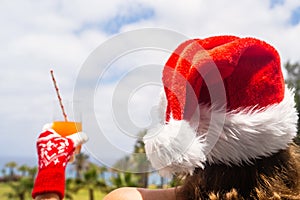 Woman hands in christmas gloves  and Santa Claus hat relaxing and drinking cocktail on balÑony opposite palm trees beach and sea