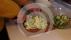 Woman hands cook, chop, slicing cabbage with knife on wooden cutting board near glass bowl with vegetables in kitchen