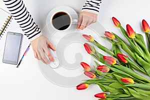 Woman hands with cup of coffee and computer mouse. Mobile phone, paper notebook and beautiful flowers on white table