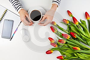 Woman hands with cup of coffee and computer mouse. Mobile phone, paper notebook and beautiful tulips on white table