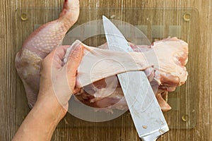 Woman hands cutting off skin from raw chicken leg with knife raw on glass cutting board on wooden background