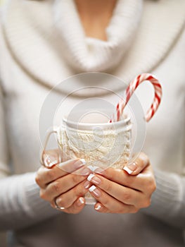 Woman hands with elegant french manicure nails design holding a cozy knitted mug with cocoa and a candy cane.