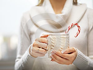 Woman hands with elegant french manicure nails design holding a cozy knitted mug with cocoa and a candy cane.