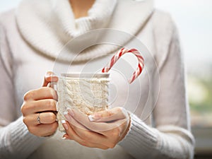 Woman hands with elegant french manicure nails design holding a cozy knitted mug with cocoa and a candy cane.