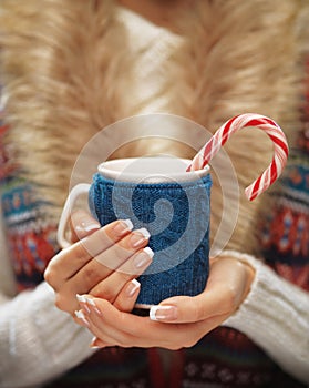 Woman hands with elegant french manicure nails design holding a cozy knitted mug with hot cocoa or coffee with a mint candy cane.