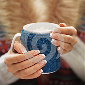 Woman hands with elegant french manicure nails design holding a cozy knitted mug. Winter and Christmas time concept.
