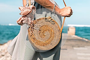 Woman hands with fashionable stylish nude rattan bag and silk scarf outside. Tropical island of Bali, Indonesia. Rattan