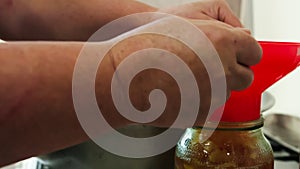 woman hands filling glass jars with apple jam via red funnel, slow motion closeup