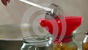 Woman hands filling glass jars with apple jam via red funnel and soup ladle, slow motion closeup